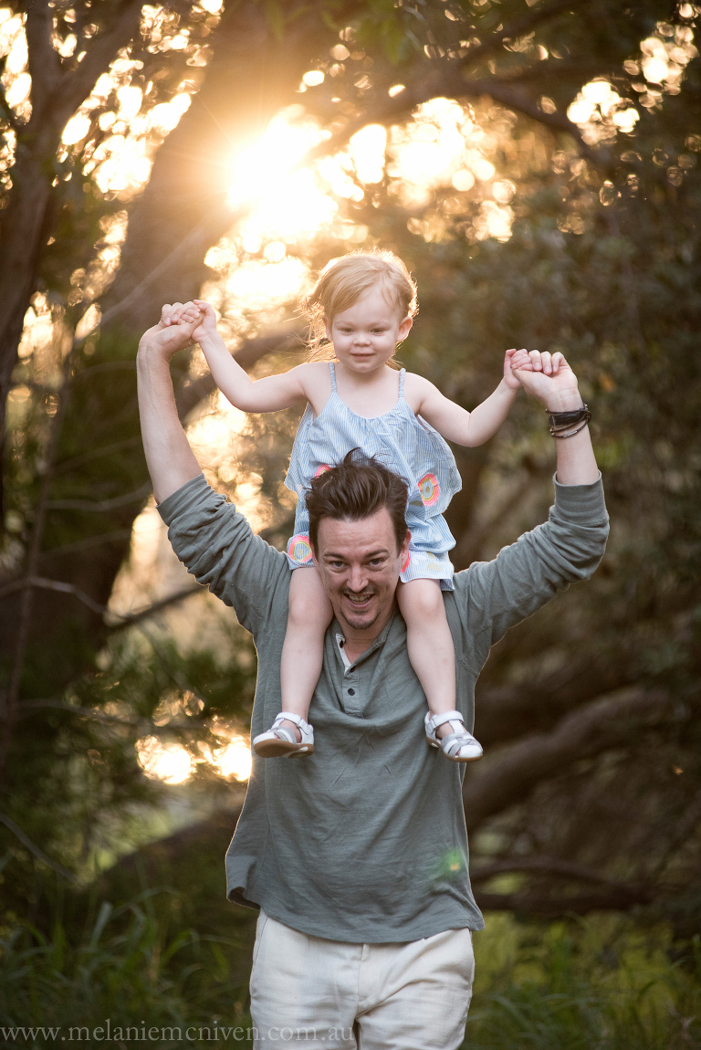 A girl on her dads back with the sun filtering through trees